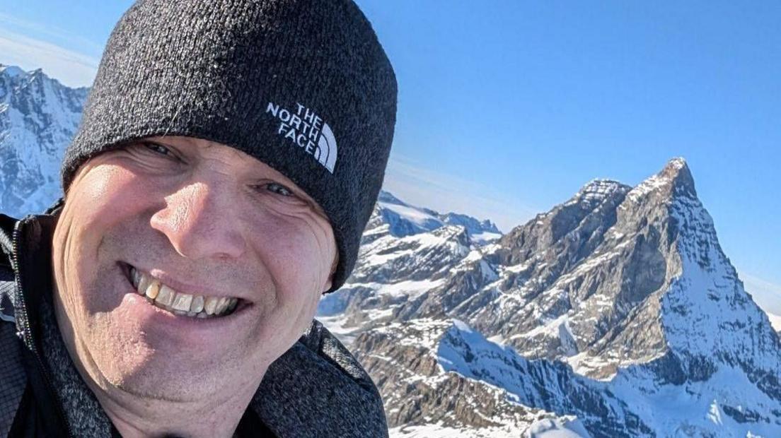 Close-up of a smiling man in a woolly hat in front of a mountain.