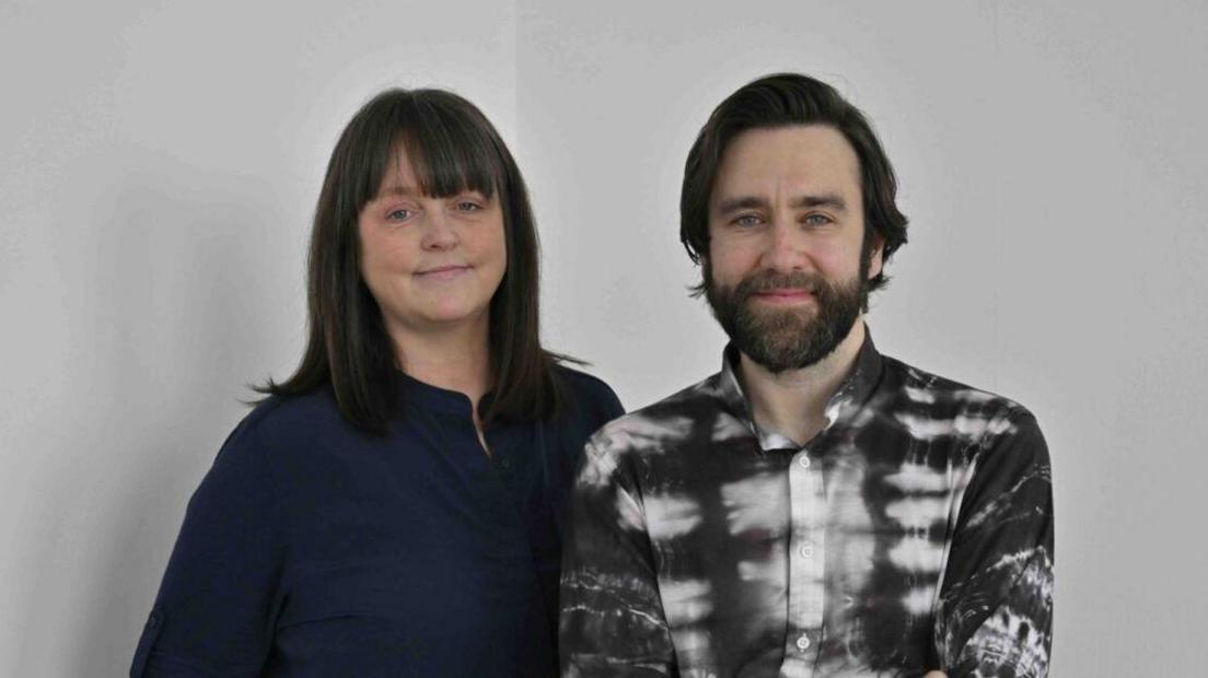 A man and woman smiling stanging in front of a white wall