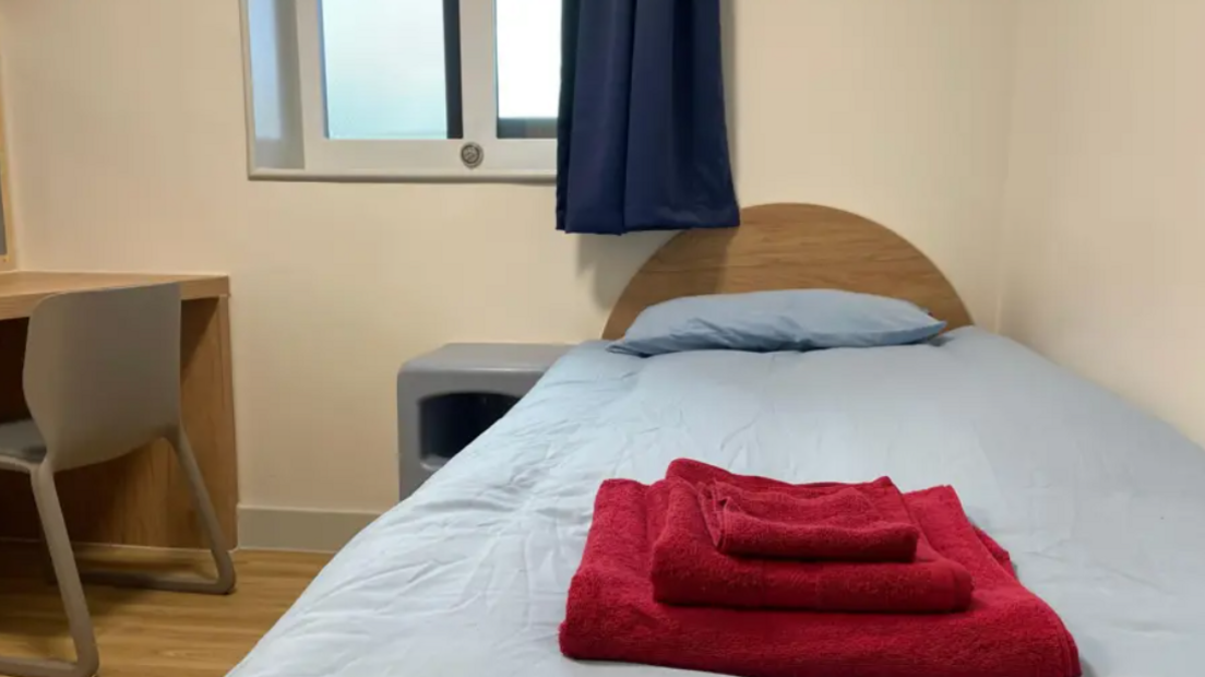 A bedroom with cream walls, a window with navy blue curtains and a wooden bed with pale blue bedding. There is a wooden desk and chair beside the bed, with a pile of red towels on the bed.