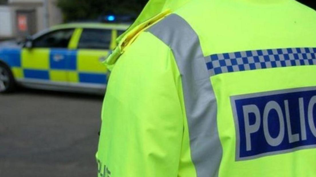 The image shows a police officer wearing a high‑visibility jacket in the foreground, with a police vehicle visible in the background.