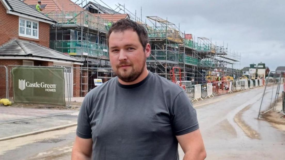 Nathan Long stand on the street in front of a houses with scaffolding and builder's fencing.