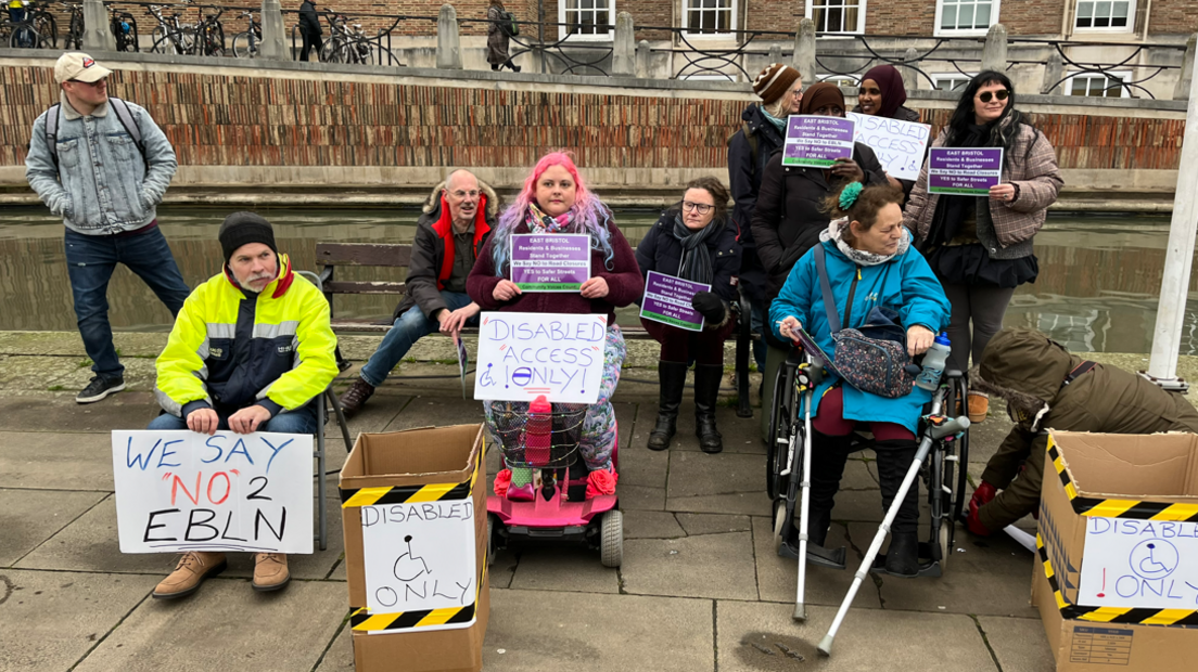 A group of protestors sit out side Bristol City Hall holding signs reading "We say 'no' to EBLN" and "Disabled access only".