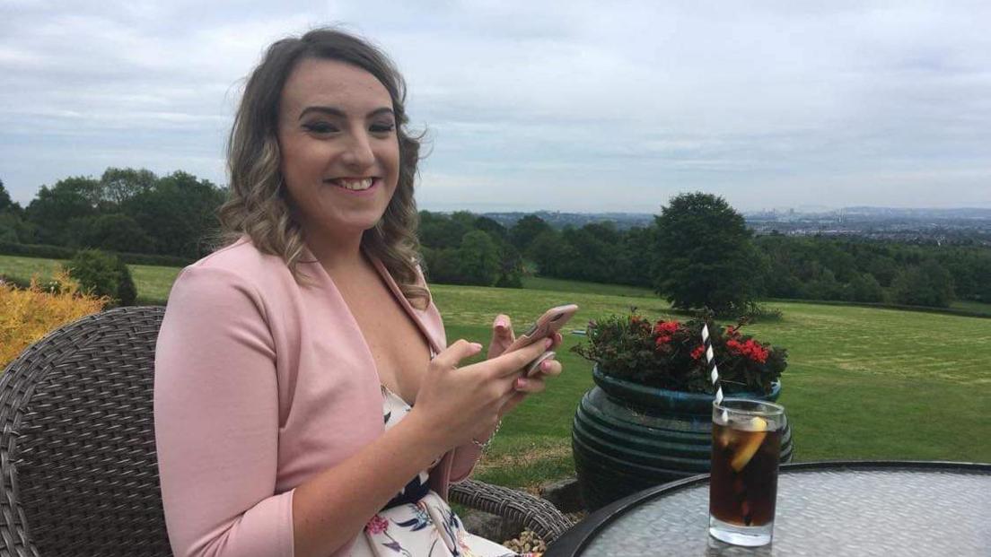 A blonde woman wearing a pink jacket over a white dress smiles at the camera. She has a drink in front of her and is sat at a table in a rural area with trees and fields behind her.