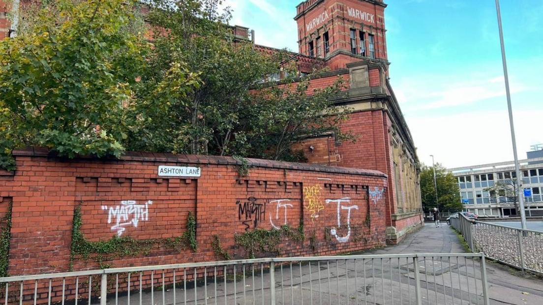 A wall by the mill covered in graffiti. The tower on the mill reads Warwick in white lettering. There is grey railing in front of the wall - which also has a sign which reads Ashton Lane.