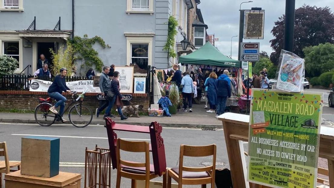 Magdalen Road Food Festival - an image taken looking down the stalls on the road from behind various types of furniture at another stall. Several people and a dog can be seen in the street. There is bunting hung around one stall under a green marquee