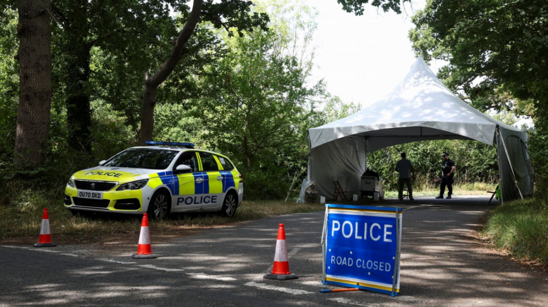 A police car and police "Road Closed" sign on a country road, with two police officers standing under a large  which gazebo which spans the road