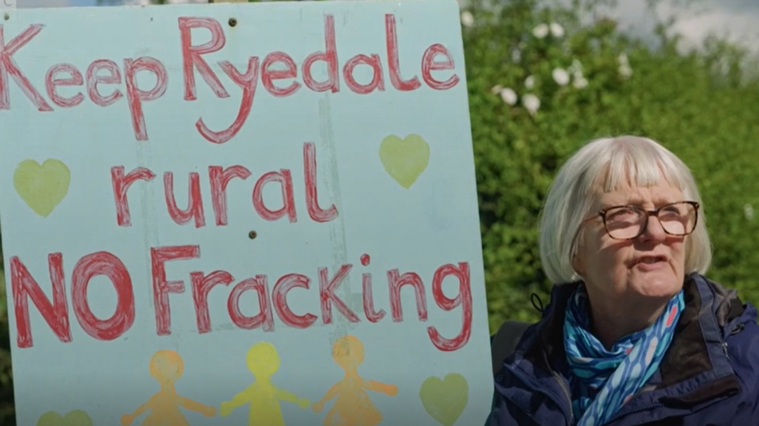 A woman with grey hair and glasses holds up a placard reading 'keep Ryedale rural - no fracking.' 