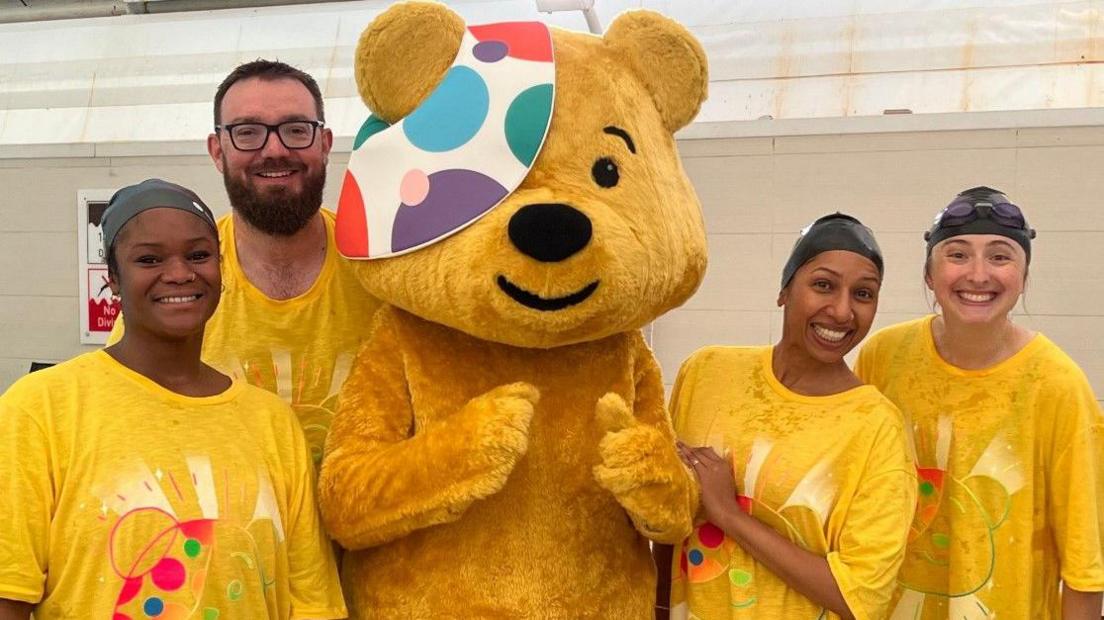 Sali Shobowale, Phil Mercer, Rena Annobil and Katherine Bett gathered around a giant Pudsey Bear at the pool. They are smiling and posing for the camera, wearing yellow t-shirts and swimming caps