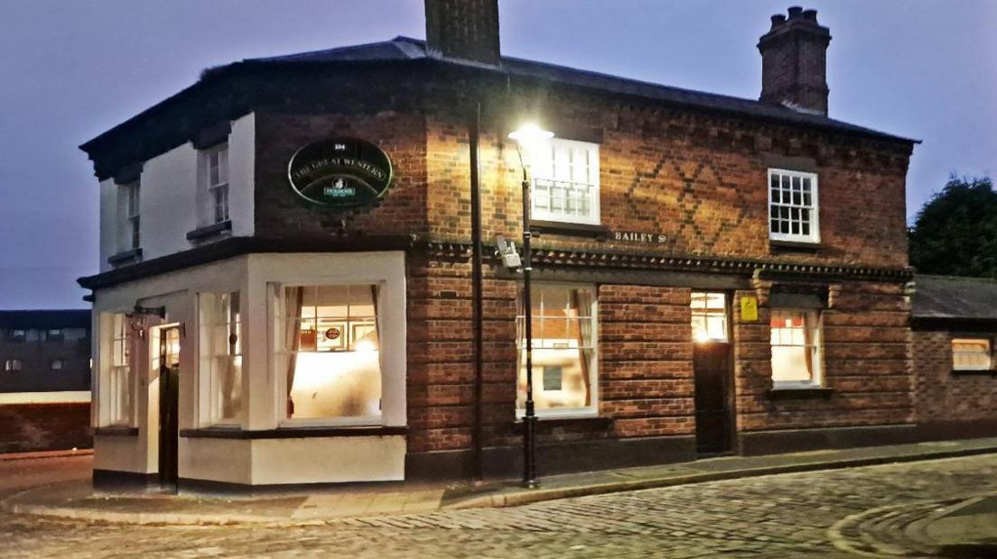 The corner of a brick building on a cobbled street in the evening with lights on in the windows and a street light standing outside the door. The building has a white front and an oval black sign on the wall