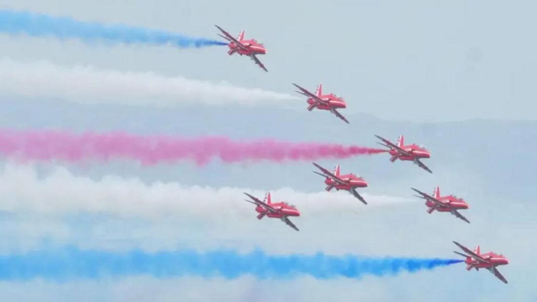 Aerial shot of seven Red Arrow jets in the sky from Airbourne 2024  in formation with red, white and blue smoke coming out of the rear of the planes.  