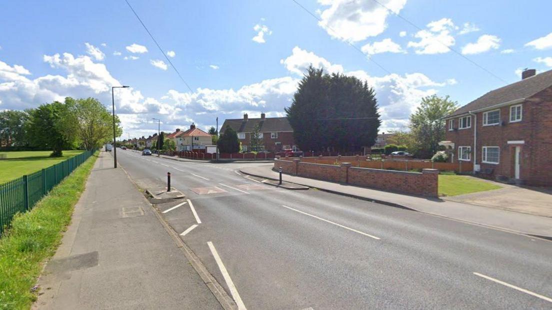 A residential street on a sunny day. Homes line the street on the right while a big green space can be seen on the left. Two speed bumps can be seen in the middle of the road.