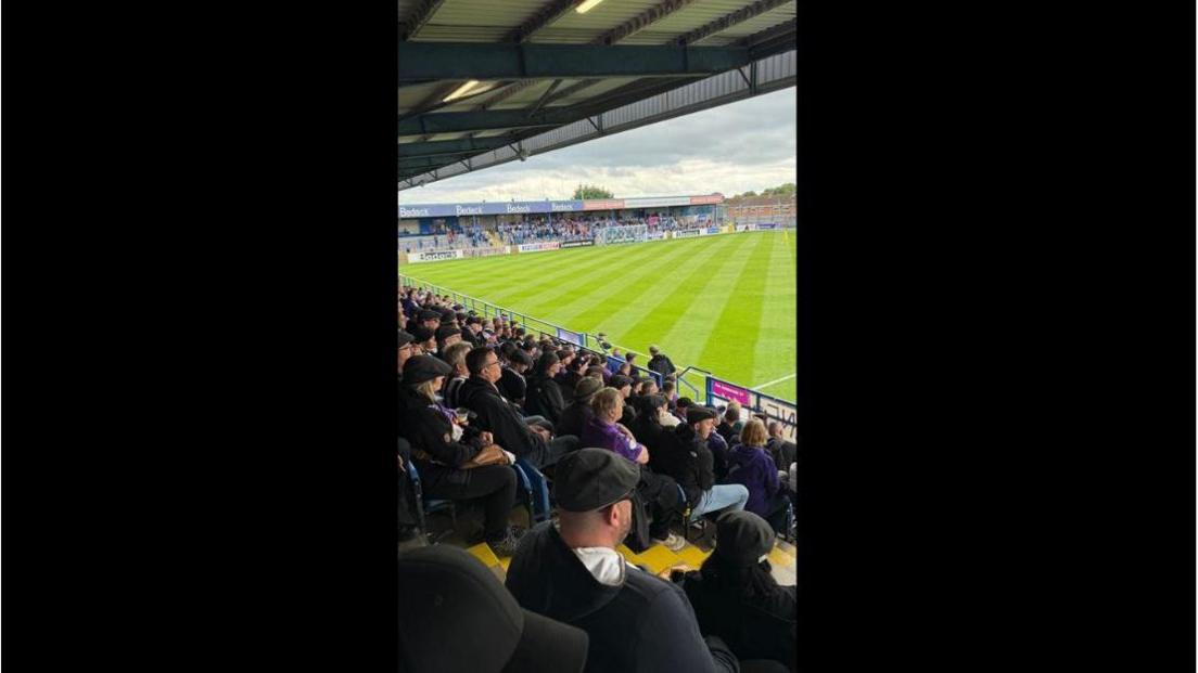 A football stadium filled with fans. The camera is pointing towards one goal end. The grass is cut in a crisscross pattern. 