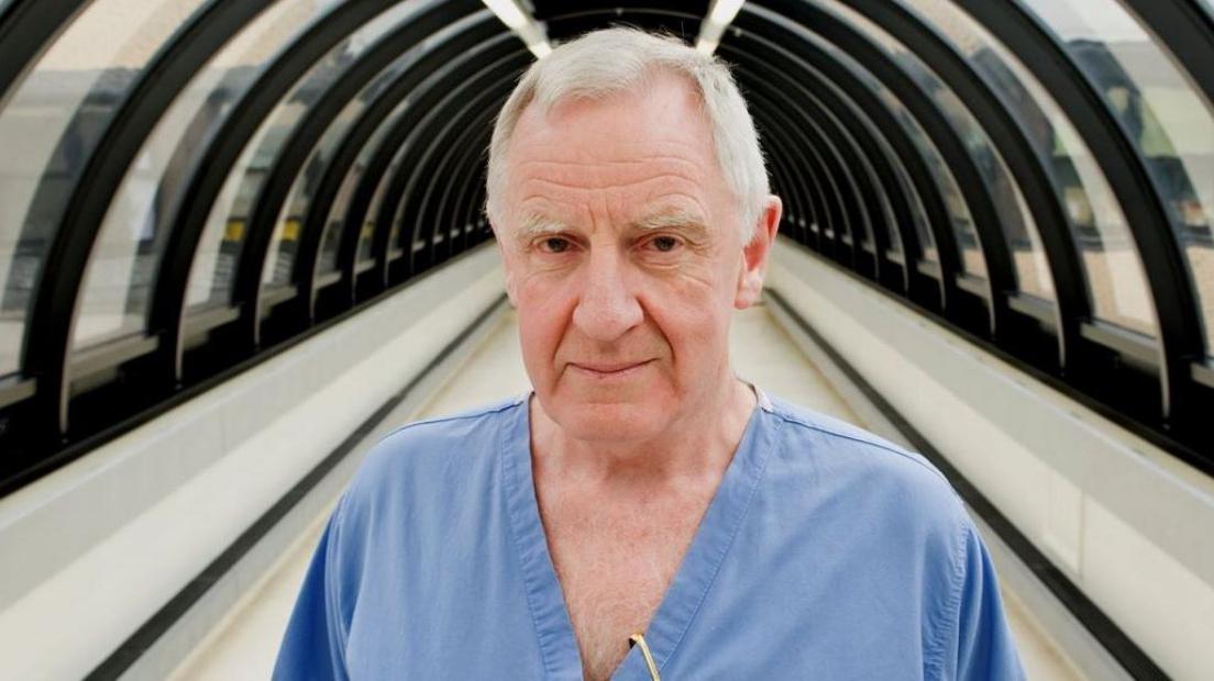 A older man with white hair wearing blue surgical scrubs stands in a covered walkway at a hospital. 