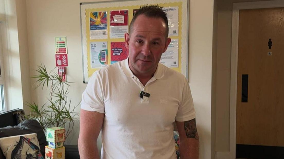 A man in a white polo-shirt is standing looking at the camera. He is looking at the camera. He has short dark hair. He is standing in front of a wall that has a notice board on it with a pot-plant and some children's toys visible.