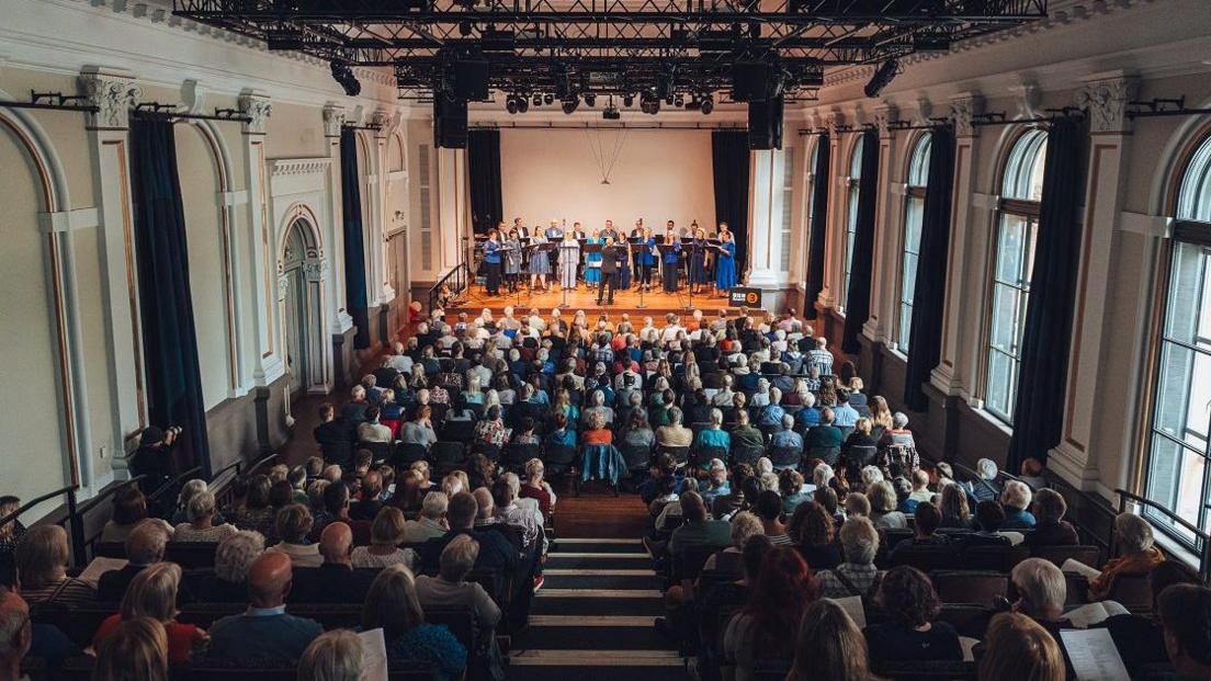 Hundreds of people are seated watching the BBC singers perform on the stage in The Lantern at Bristol Beacon. The photograph is taken from the back of the auditorium