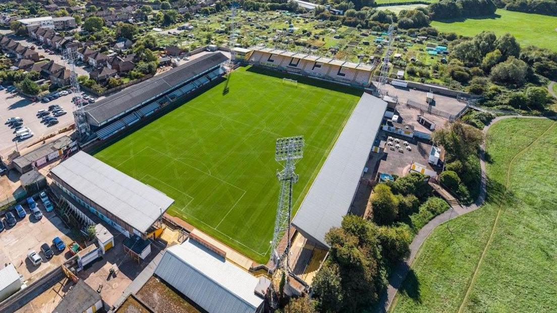 Aerial shot of Cambridge United's Abbey Stadium, showing stands on four stands, four tall light towers at each corner, allotments beyond to its back and a park to its right