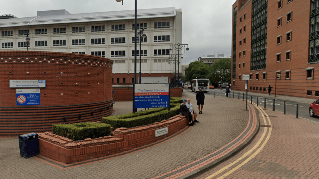 Blue-and-white entrance sign to Leeds General Infirmary, as seen from Clarendon Way. In the background are hospital buildings and a bus, two people sit on a wall in the foreground.