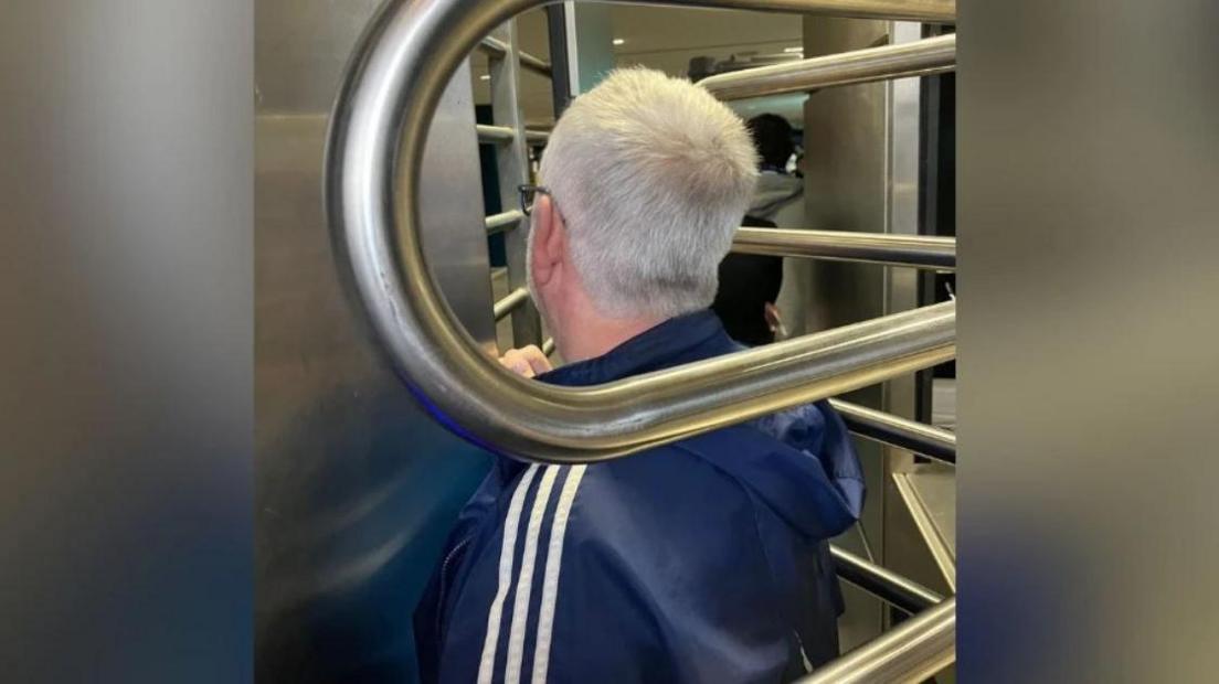 A man with grey hair faces away from the camera and appears trapped in a metal turnstile.