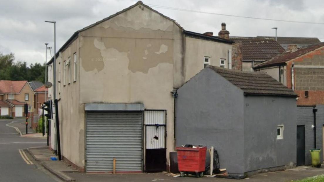 Streetview of a two-storey building with a white door with a black gate across it and a silver metal garage door. It is poorly kept with large areas of the cream rendering having come off to reveal dirty walls beneath.