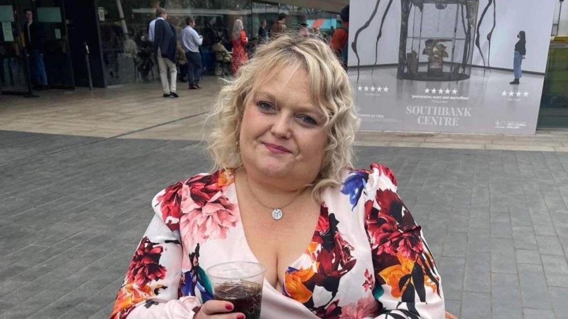 A woman with blonde hair and wearing a dress with a floral pattern is holding a drink.