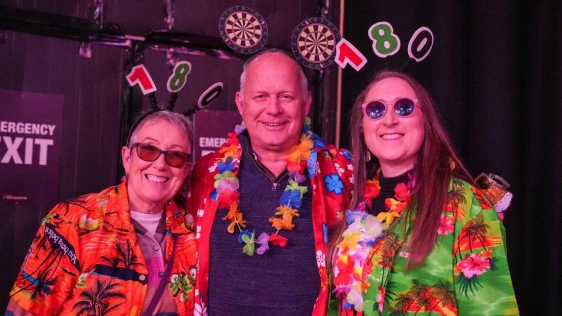 Three people in colourful tops are looking at the camera. The two on the left and right have the number 180 above their head. The man in the middle has two darts boards above his head.