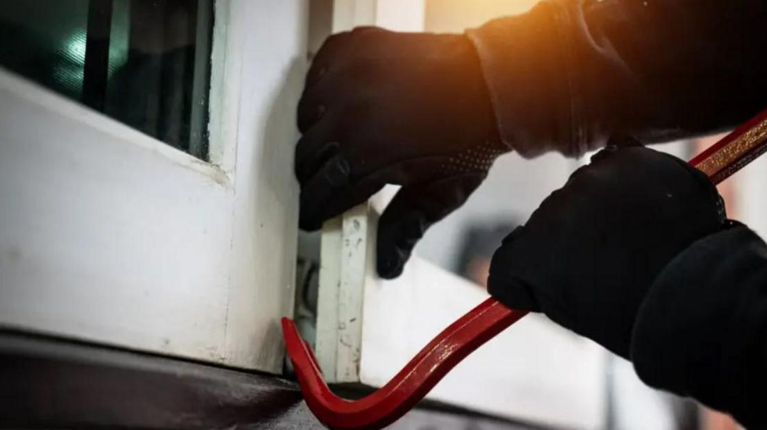 Hands in black gloves are using a crowbar to open a window