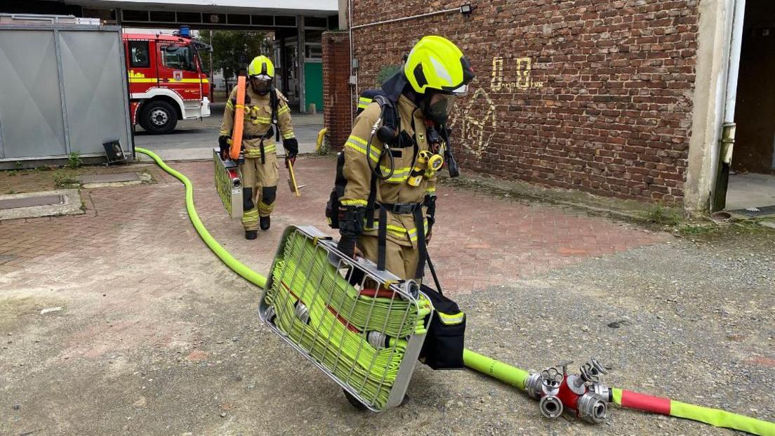 Two German firefighters in all of their protective equipment carrying a long water hose