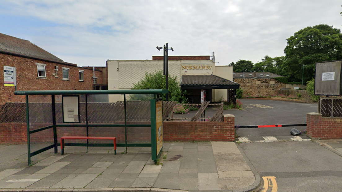 Normanby hotel is a single-storey white building with a gold sign on the front which reads: "Normanby". A small car park surrounds it, and a green bus stop is seen on the pavement out front.