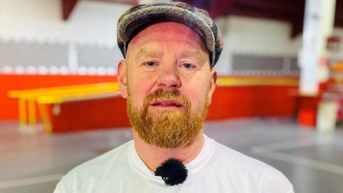 Man with ginger beard, wearing a bunnet, a white t-shirt and looking at camera.