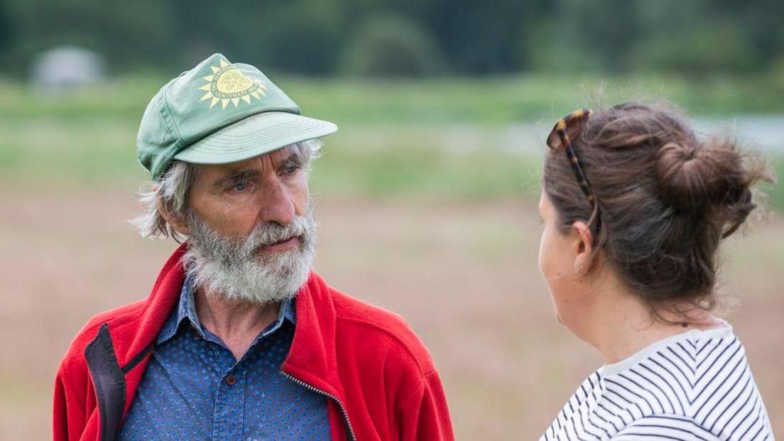 Angus Wainwright speaks with a woman at Sutton Hoo. He wears a green hat and has grey hair with a grey beard. He is wearing a red jumper with a blue shirt underneath. The woman looks away from the camera and has dark brown hair and wears a white top with blue stripes.