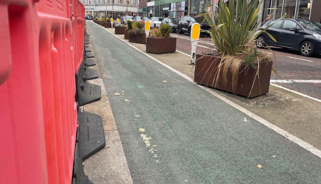 Red barriers beside a cycle lane in Belfast city centre. Large potted plants and parked cars are also visible in the picture