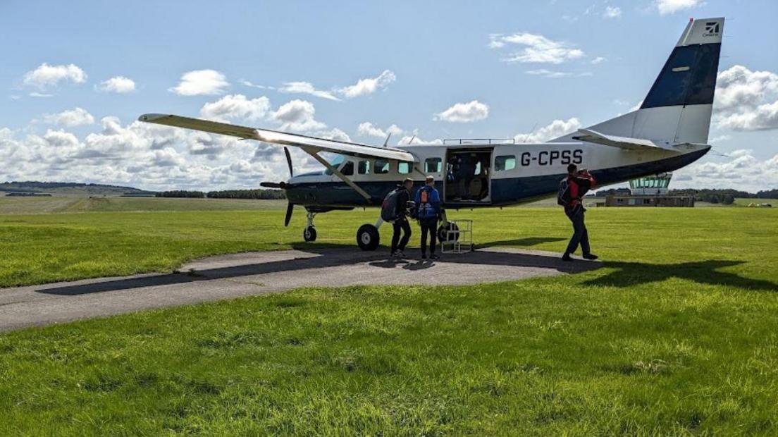 A plane parked on a tarmac patch on Netheravon Airfield as three men wearing parachutes get in. 