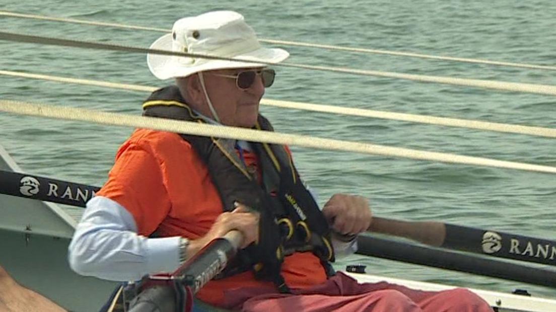 A man wearing orange sitting in a boat holding two oars. He is wearing sunglasses and a white hat.