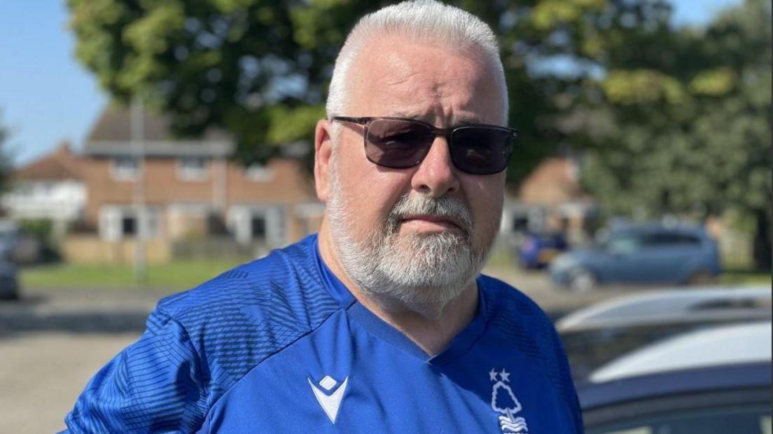 David Anscombe has short white hair and a close-cropped beard, his is wearing sunglasses and a bright blue Nottingham Forest football shirt. 