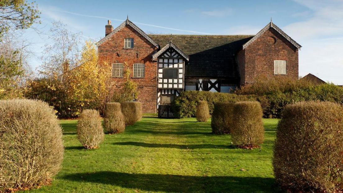 A medieval-style building can been seen, from the end of a garden walkway. The walkway is grassy with neatly trimmed bushes along each side. At the end is the hall, made both of red brick and traditional black and white Tudor architecture.