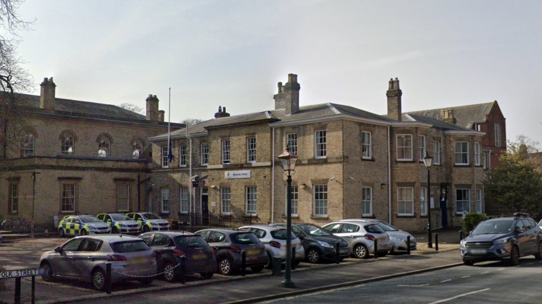 A view of a yellow brick building with a sign which reads "Beverley Police Station". A number of cars are parked outside including three police vehicles.