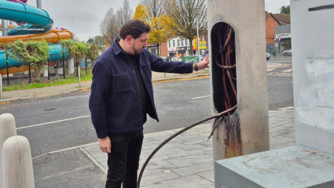 A man is standing next to a damaged mast. A leisure centre and other businesses and shops are in the background. The door of the mast is open and the wires inside have been burnt.