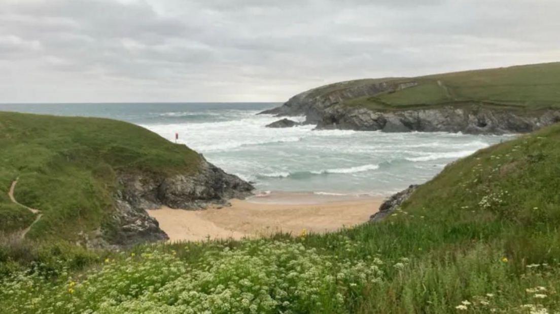 The beach at Polly Joke is a long golden stretch of sand with cliffs surrounding it. The sea can be seen in the distance with grass towards the front.
