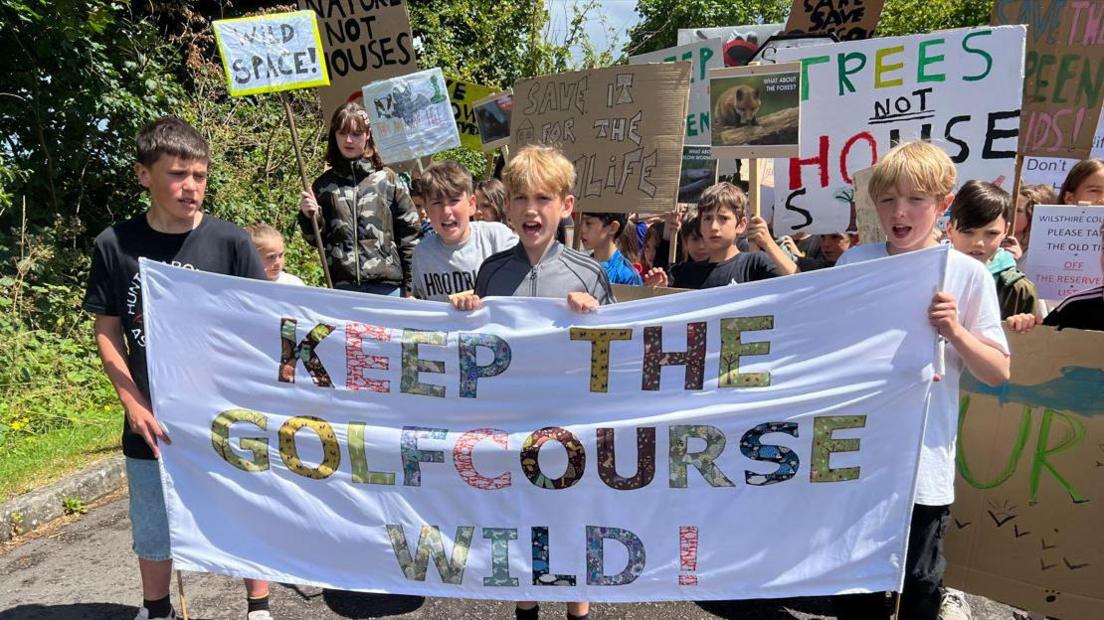 A group of schoolchildren holding a flag with multi-coloured lettering reading 'keep the golf course wild!'.