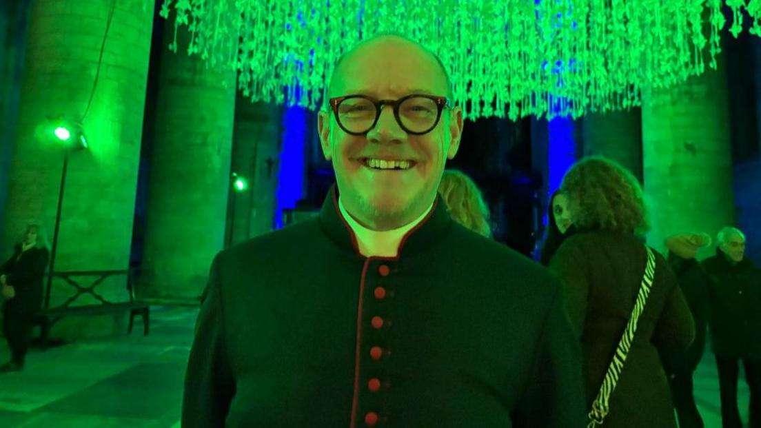 Thousands of paper doves hung from the ceiling of an abbey with green lighting shining over them and Chris Milton standing below them smiling at the camera.