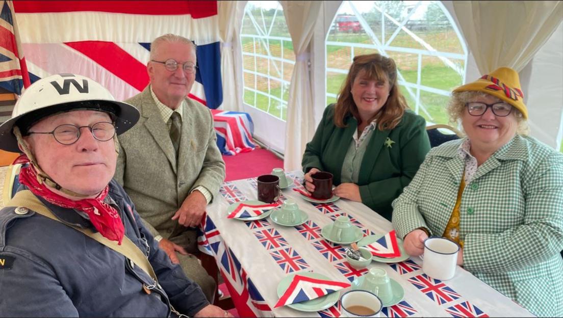 Four people sitting around a table wearing 1940s clothing. They are smiling to the camera and on the table are flags of Great Britain. They are sitting inside a marquee.
