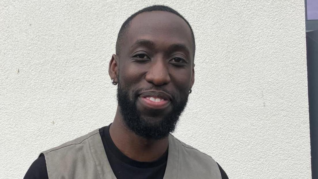 A man in a black t shirt and grey waistcoat stands in front of a grey wall. he has short black hair and beard and is smiling