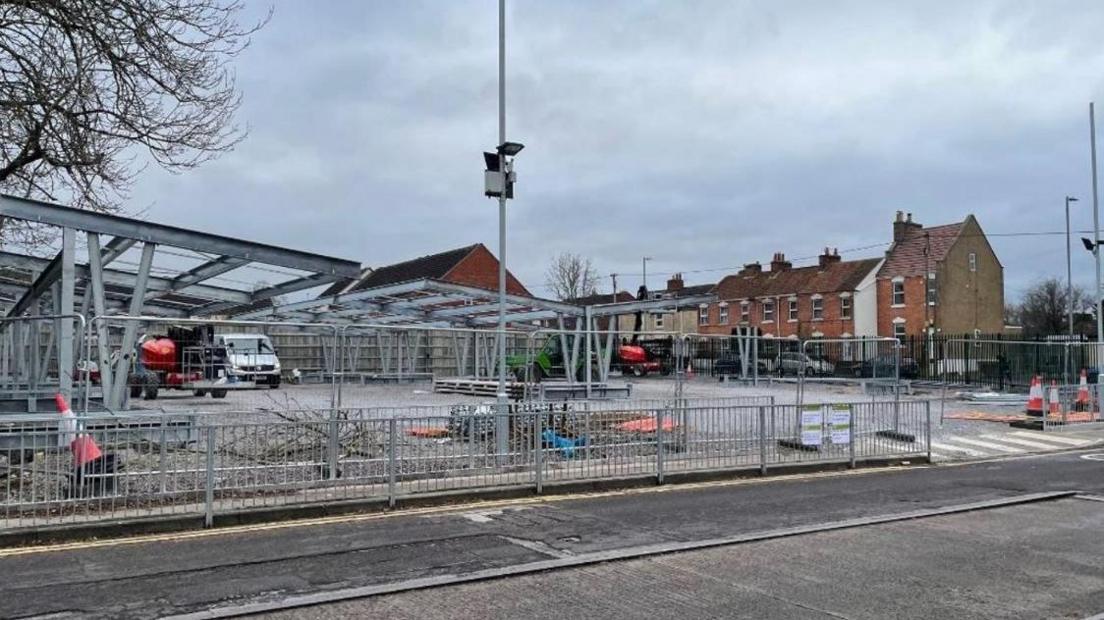 A large construction site where metal frames are being erected. The site is surrounded by a metal security fence. A row of redbrick houses is visible in the background.