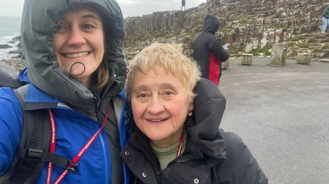 Cat Rubens and Jane Rubens stand in front a rocky area by the sea. They are both wearing warm jackets.