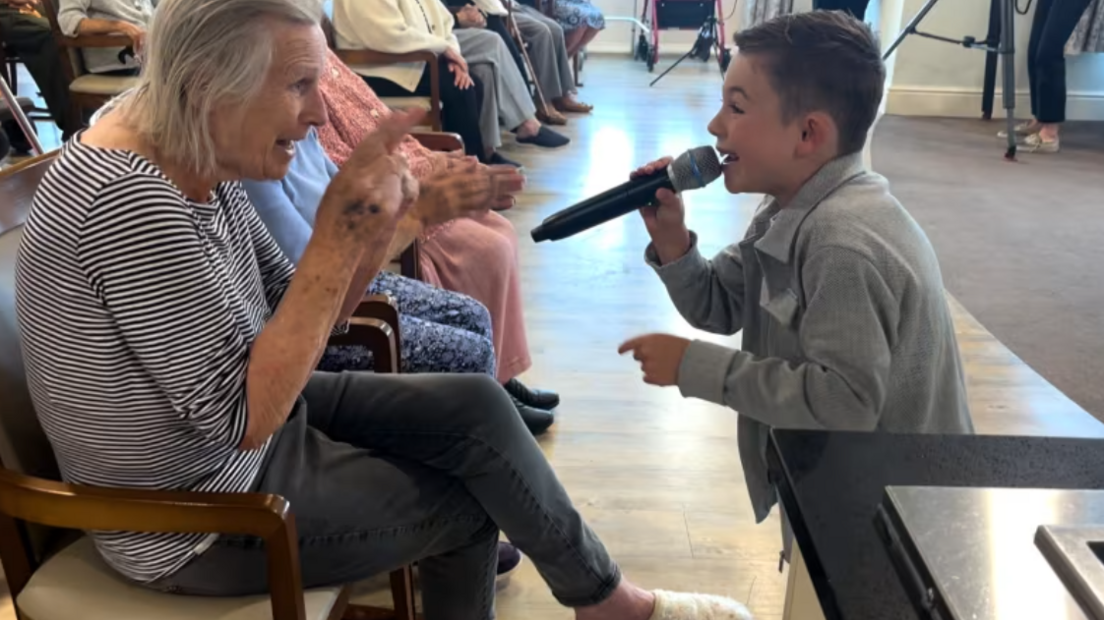 Lucciano singing on his knees with a microphone before an elderly care home resident