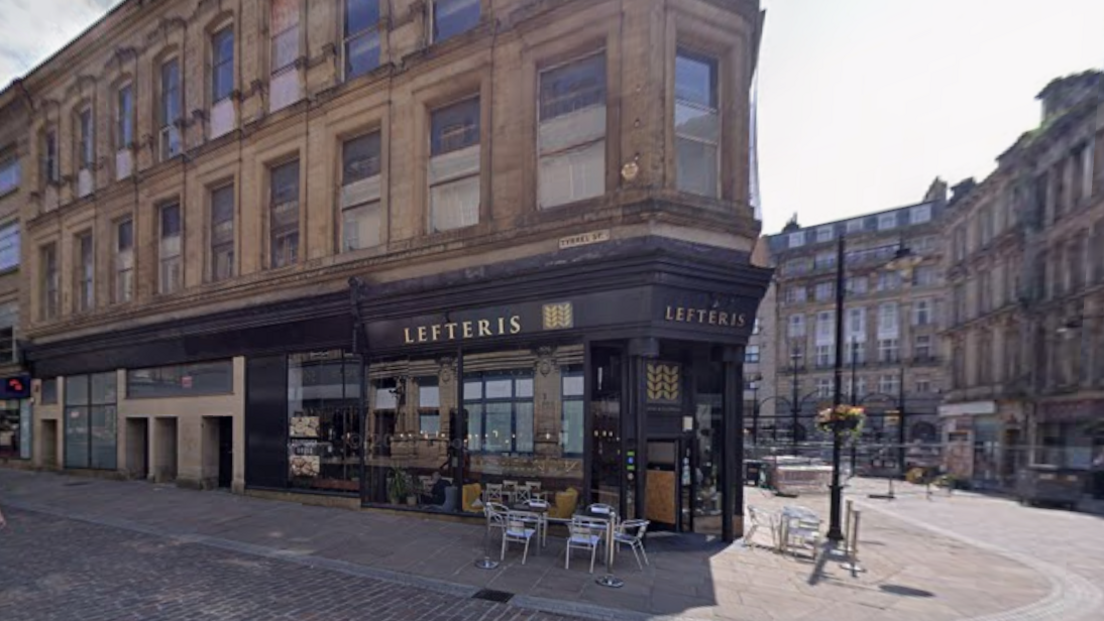 A corner of the former Brown, Muff & Co department store building. The ground floor has a black façade, and there are some metal tables and chairs outside. There are two floors with windows above. The area is surrounded by paving.