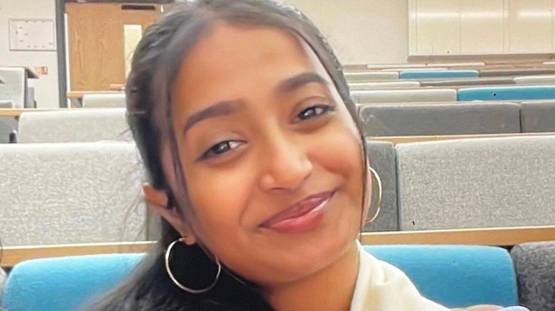 Ann Sabu Is smiling at the camera while taking a photo of herself in a lecture theatre. She has dark hair and wears a white scarf with a blue top alongside some gold hoop earrings. She rows of padded seats behind her are blue, grey and a lighter shade of grey. 