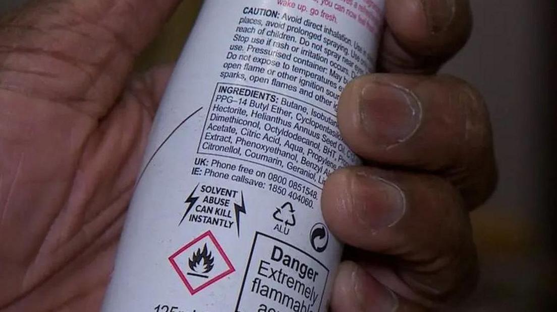 A hand holds the back of a white can of deodorant showing the ingredients and warnings about the contents.