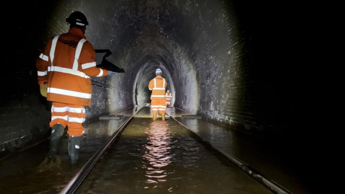 Four men in orange protective clothing, helmets and torches around their heads working inside Bransty Tunnel at Whitehaven. The tunnel floor is flooded.