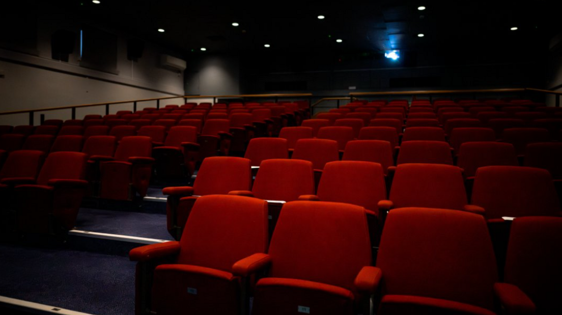 A dark auditorium with tiers of seats covered in red fabric. There is a blue-carpeted aisle down the middle. Spotlights can be seen in the ceiling.